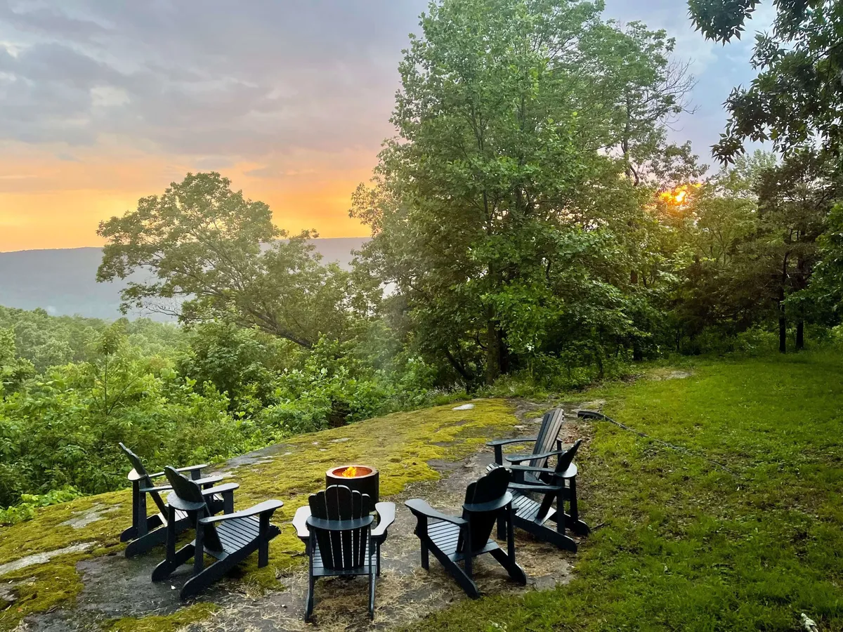 A serene outdoor fire pit area surrounded by Adirondack chairs, set against a colorful sunset and encircled by tall trees. The scene invites conversation and warmth, highlighting the opportunity for evening gatherings in the heart of nature.
