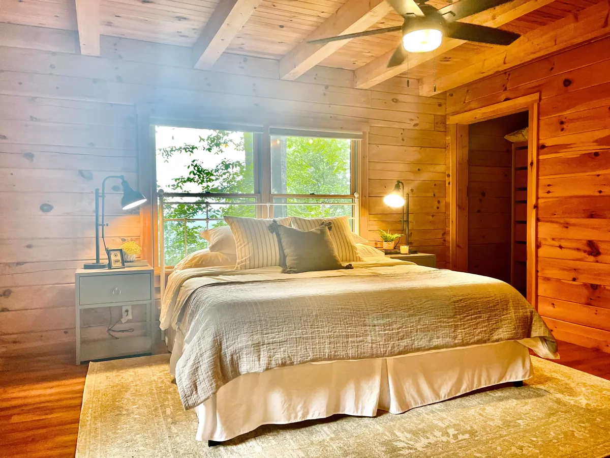 A tranquil bedroom featuring a cozy bed with premium linens, a nightstand with a lamp, and large windows that let in abundant natural light with treetop views. The wood-accented decor and ceiling fan create a restful, nature-inspired ambiance.