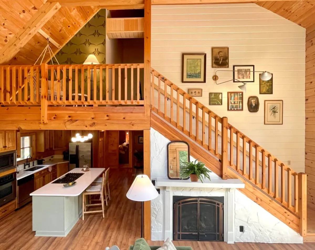 A spacious open-plan living area featuring a prominent staircase, a cozy gas fireplace with a beautifully designed mantle, a kitchen island at the edge of the frame, and carefully curated wall art and decor. The inviting living room lamp and ample seating further enhance the welcoming vibe.