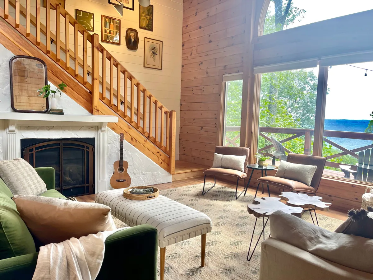 A warm, sunlit living room with a green sofa, large windows providing views of the surrounding greenery, a staircase, a gas fireplace, and an art-adorned wall. The room feels both inviting and filled with creative energy.
