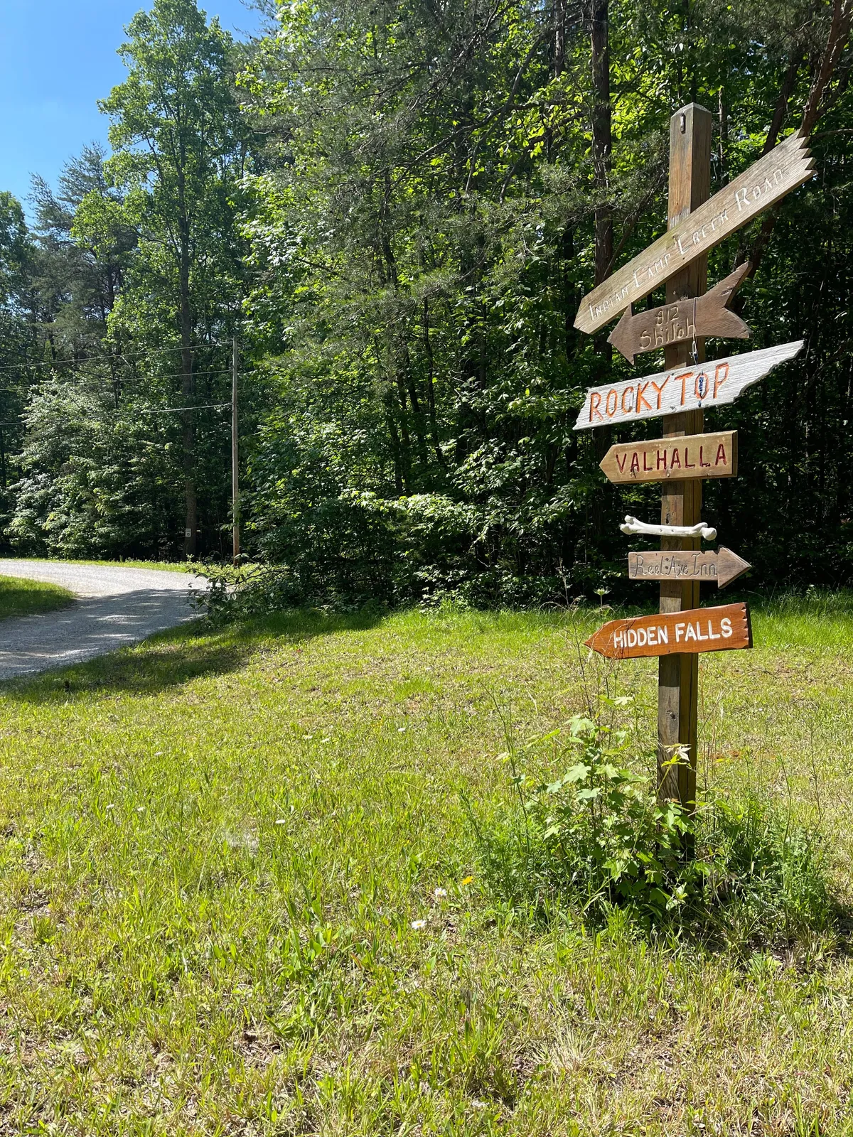 A wooden directional sign emerges from lush greenery along a gravel pathway, signaling the start of a hiking trail into the expansive wooded preserve. The setting is peaceful, hinting at the vast network of trails and hidden nature spots awaiting discovery.
