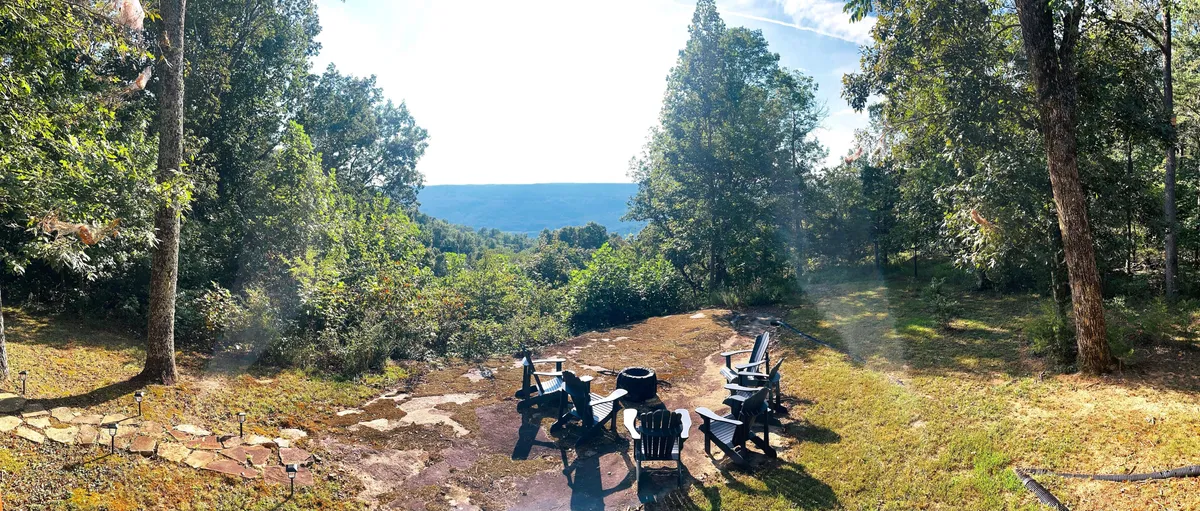 An exterior scene featuring a cozy fire pit surrounded by Adirondack chairs, with a backdrop of mountains and trees. The setting is designed for social gatherings, relaxation, and evenings under the stars.