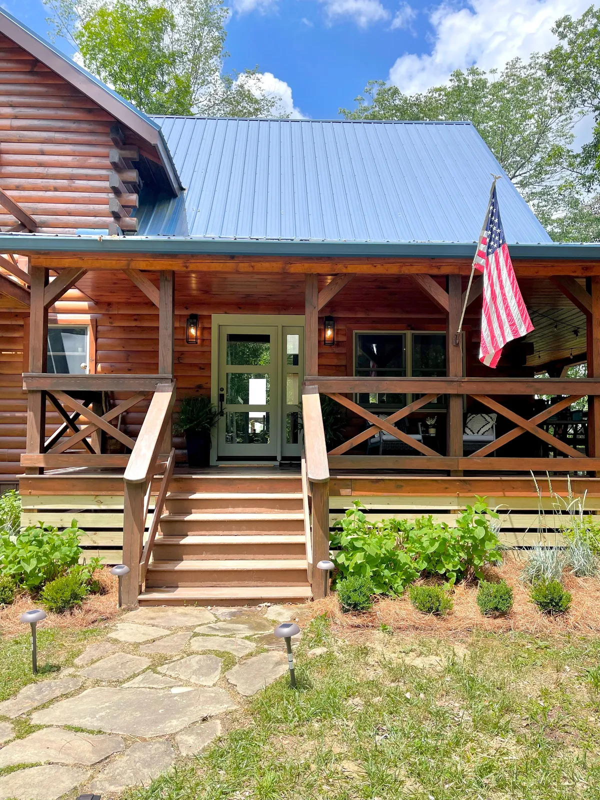 This image captures the exterior of the property, featuring a welcoming front porch with steps leading up to the entrance, an American flag, and a landscaped garden. The scene suggests a space where guests can gather, relax, and enjoy the outdoors, creating a sense of arrival and connection to the local environment.