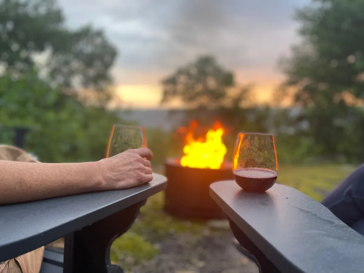 This exterior image centers on a fire pit surrounded by glasses of wine and the hands of individuals gathered together. The background suggests a peaceful outdoor setting, ideal for winding down after a day of adventure.