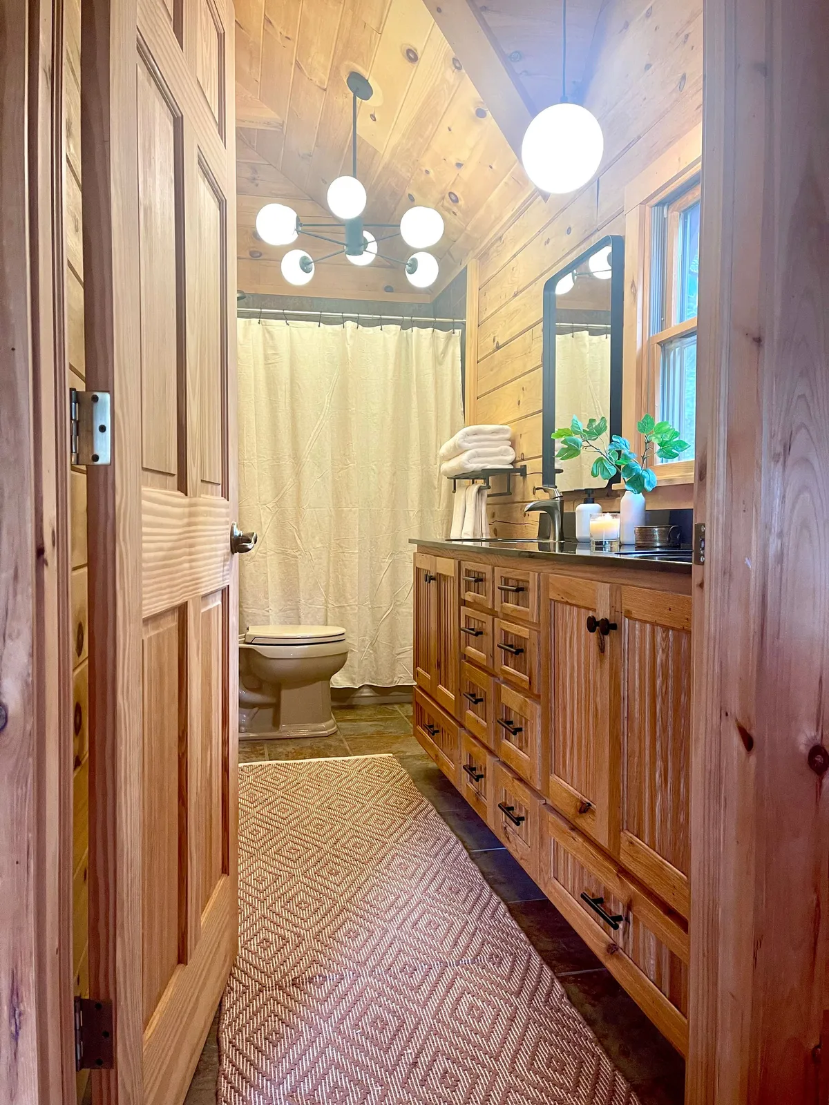 A modern bathroom with wooden cabinetry, sleek light fixtures, a vanity mirror, a shower curtain, decorative towels, and a green plant on the counter. The ambiance is serene, offering a restorative setting for self-care routines.