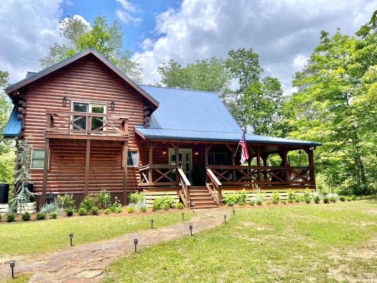 This image showcases the exterior of the Mossy Falls Cabin, highlighting its log cabin structure, blue metal roof, front porch, and American flag. The setting exudes a welcoming and rustic charm, surrounded by nature and inviting relaxation on the porch.