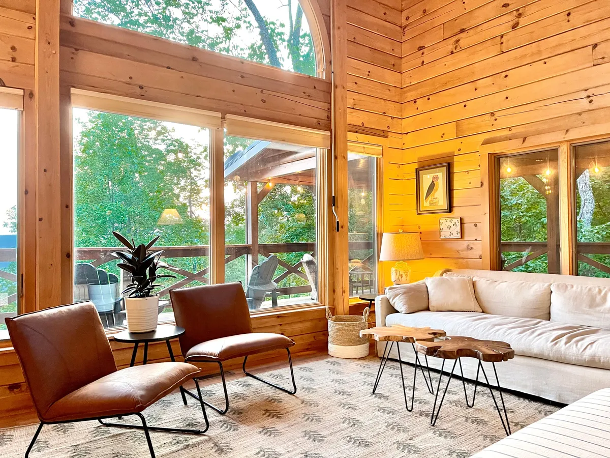 A luminous living area featuring large windows that frame lush greenery, a comfortable seating arrangement, and stylish decorative accents. The scene is inviting, with natural light filling the space and a sense of calmness. The environment suggests relaxation and socializing, enhanced by the view to the outside world.