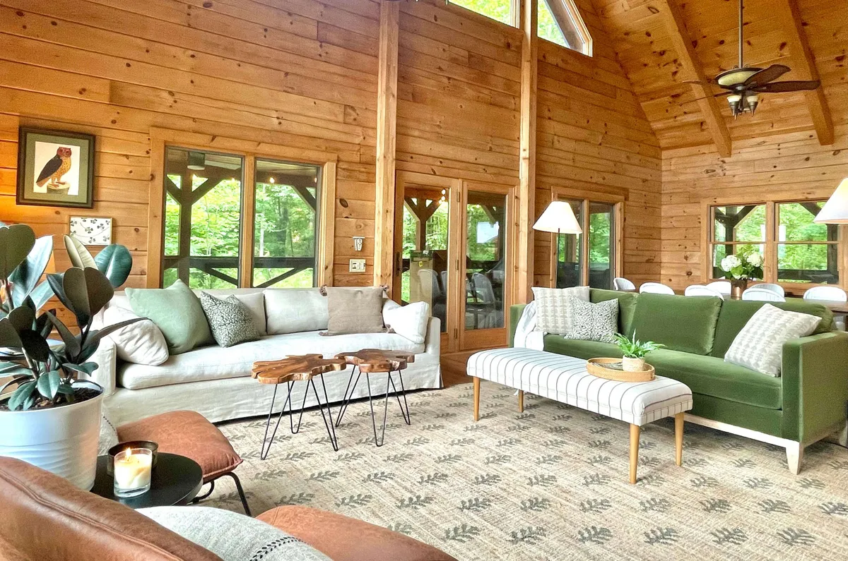 Inside, the cabin’s living area features elegant wooden paneling, large windows framing forest views, a green sofa, striped ottoman, and artful decor. The space invites relaxation, reading, and intimate conversation in a plush, thoughtfully designed setting.
