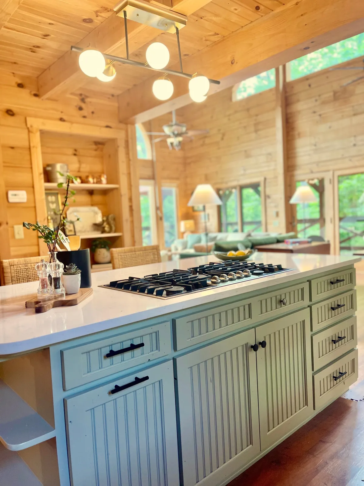 A modern, fully equipped kitchen with a gas cooktop on a central island, open shelving, warm wooden cabinets, and striking light fixtures. The setup is clearly designed for both culinary activity and communal enjoyment.