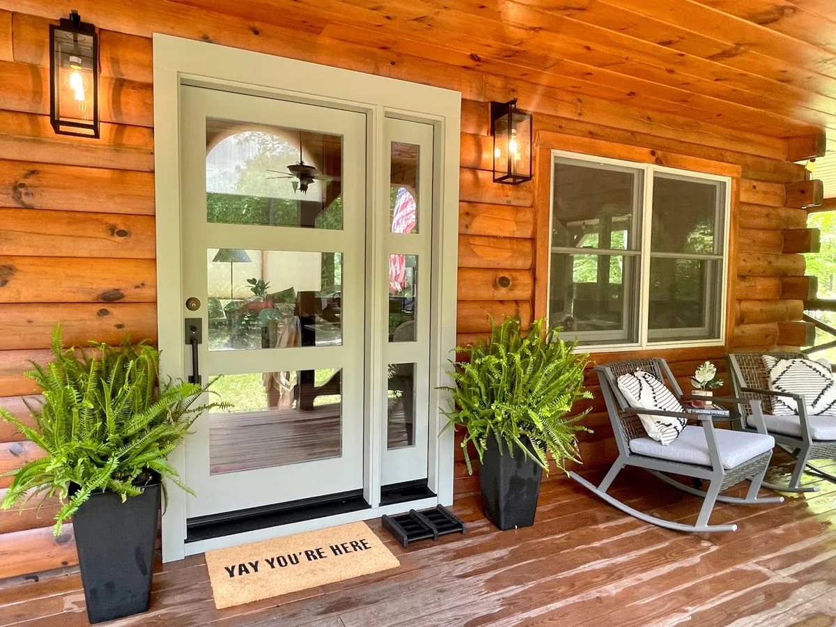 This image captures the front porch of Mossy Falls Cabin, complete with rocking chairs, a welcome mat, and planters, all inviting guests to relax and immerse themselves in the surrounding natural beauty. The porch is set against the backdrop of lush woods, providing a sense of tranquility and connection with nature.