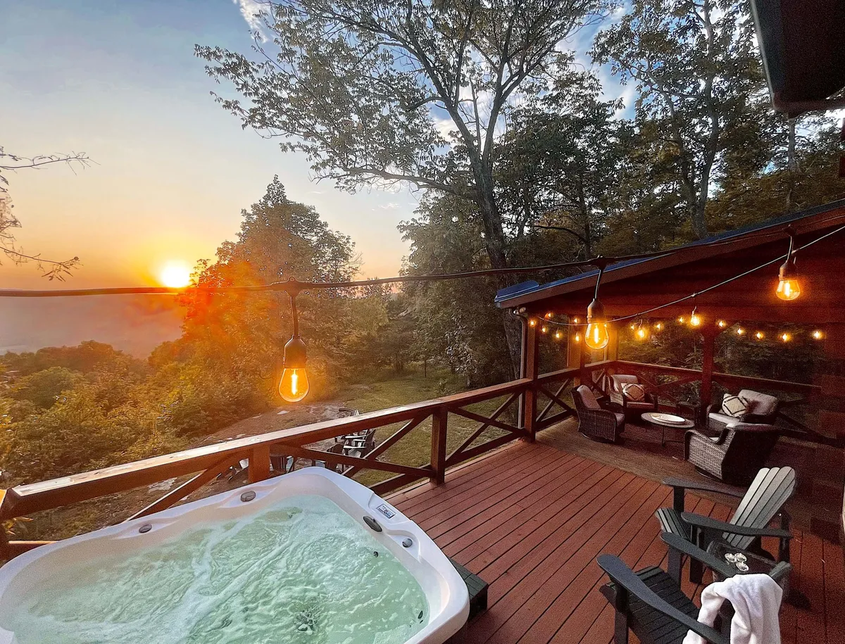 A private deck at sunset, featuring a bubbling jacuzzi, hanging lights overhead, and a cozy seating area. The view stretches out over ancient woods, creating a tranquil and luxurious escape. Guests are shown soaking in the hot tub or relaxing with drinks while enjoying the panoramic sunset.