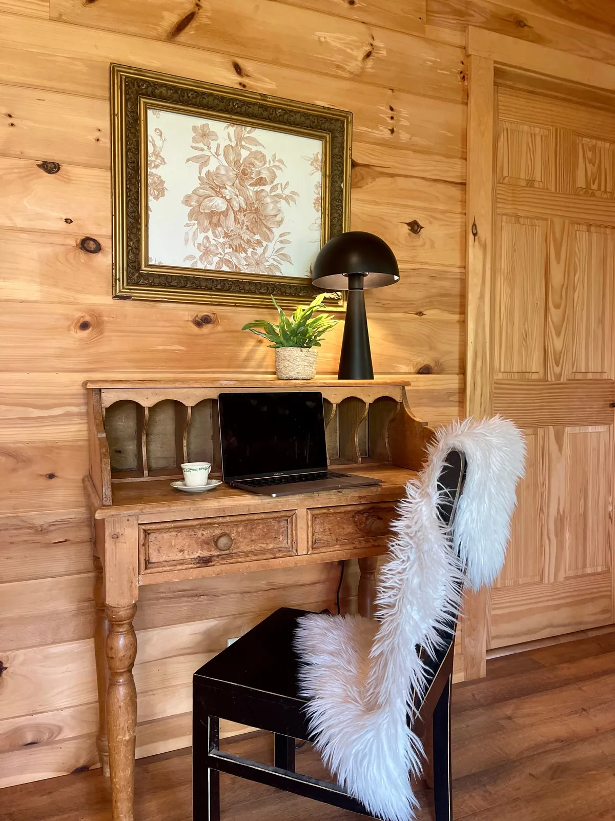 A dedicated workspace nook featuring a wooden desk, black lamp, a laptop, floral artwork, a fluffy chair cover, and a green plant. The space feels productive yet calm, perfect for creative solitude or remote work.
